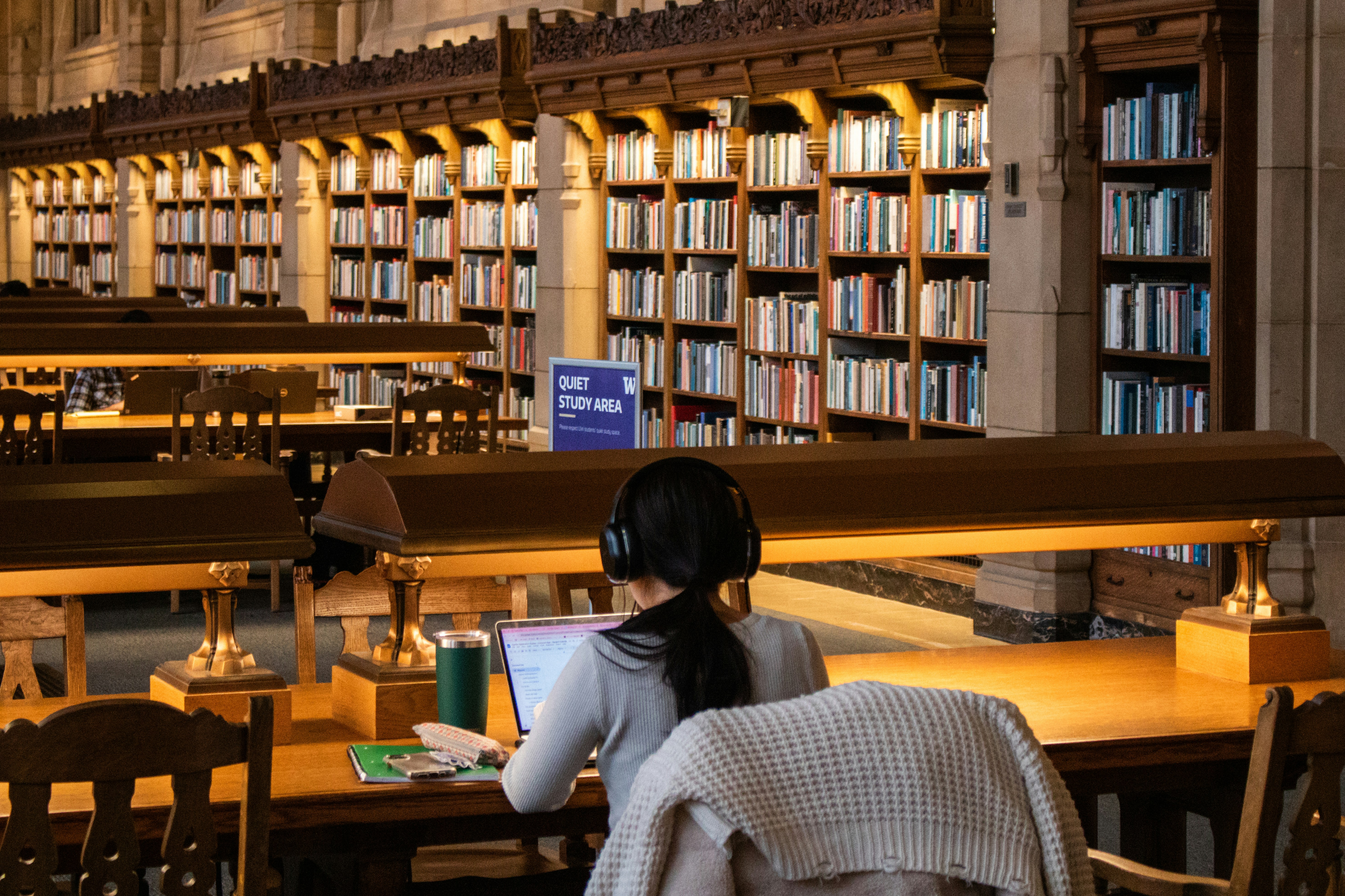 Woman with headphones on studies in a quiet library area with shelves of books in the background image